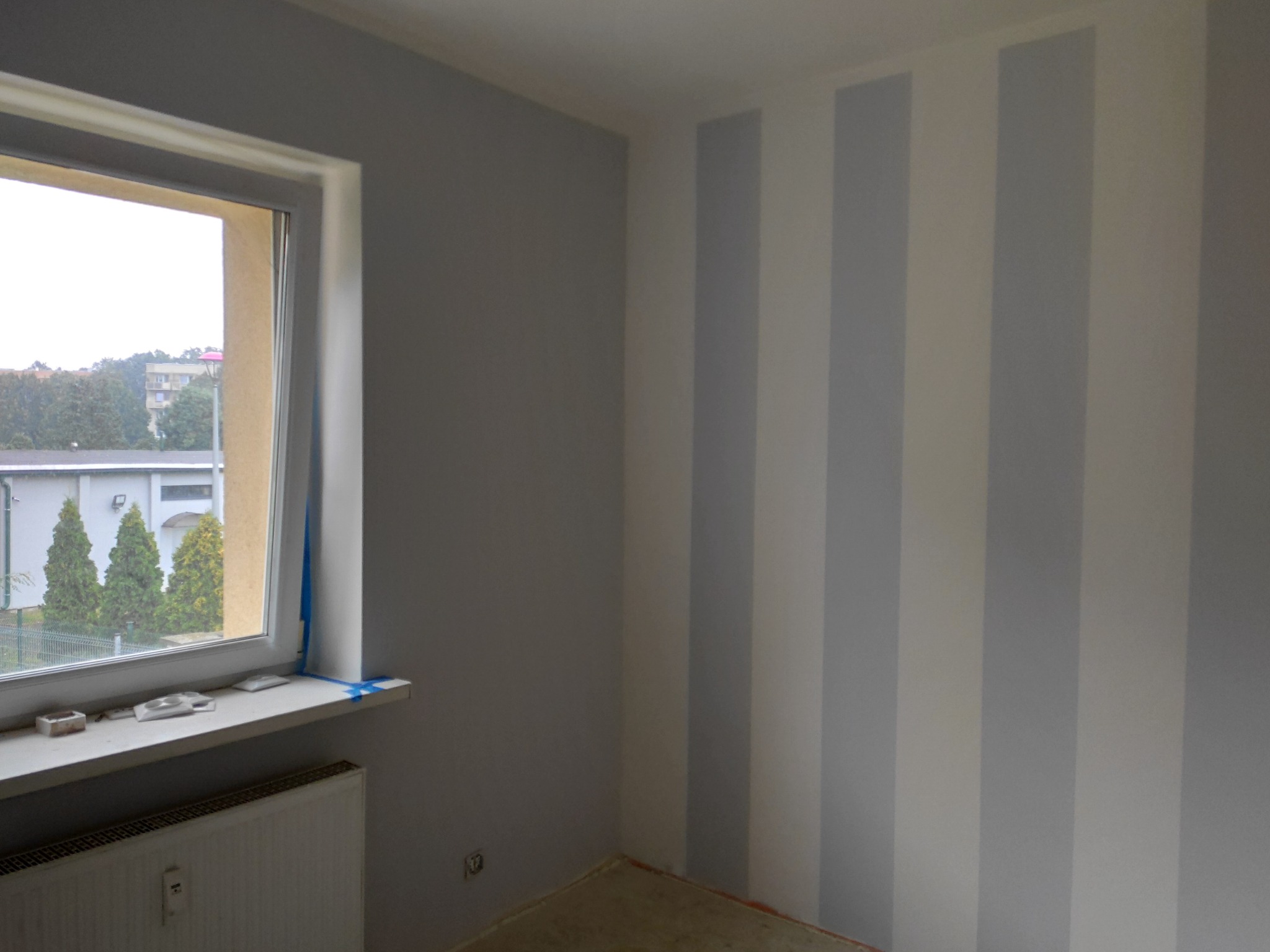Pokój po świeżym odmalowaniu w Opole, ściana w pionowe pasy szarości i bieli, okno z widokiem na zieleń, parapet z akcesoriami malarskimi zabezpieczony taśmą.