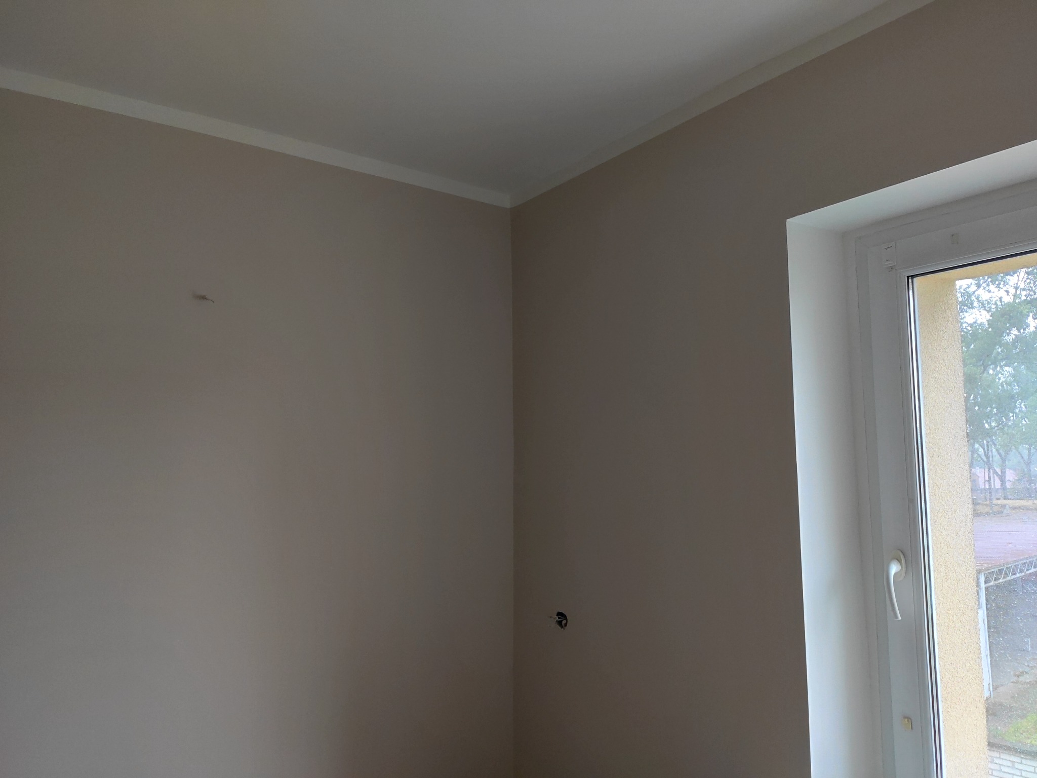 Świeżo pomalowany róg pokoju z beżowymi ścianami, białą listwą przypodłogową i oknem z widokiem na zewnątrz w pochmurny dzień. Widoczne gniazdko elektryczne bez osprzętu.