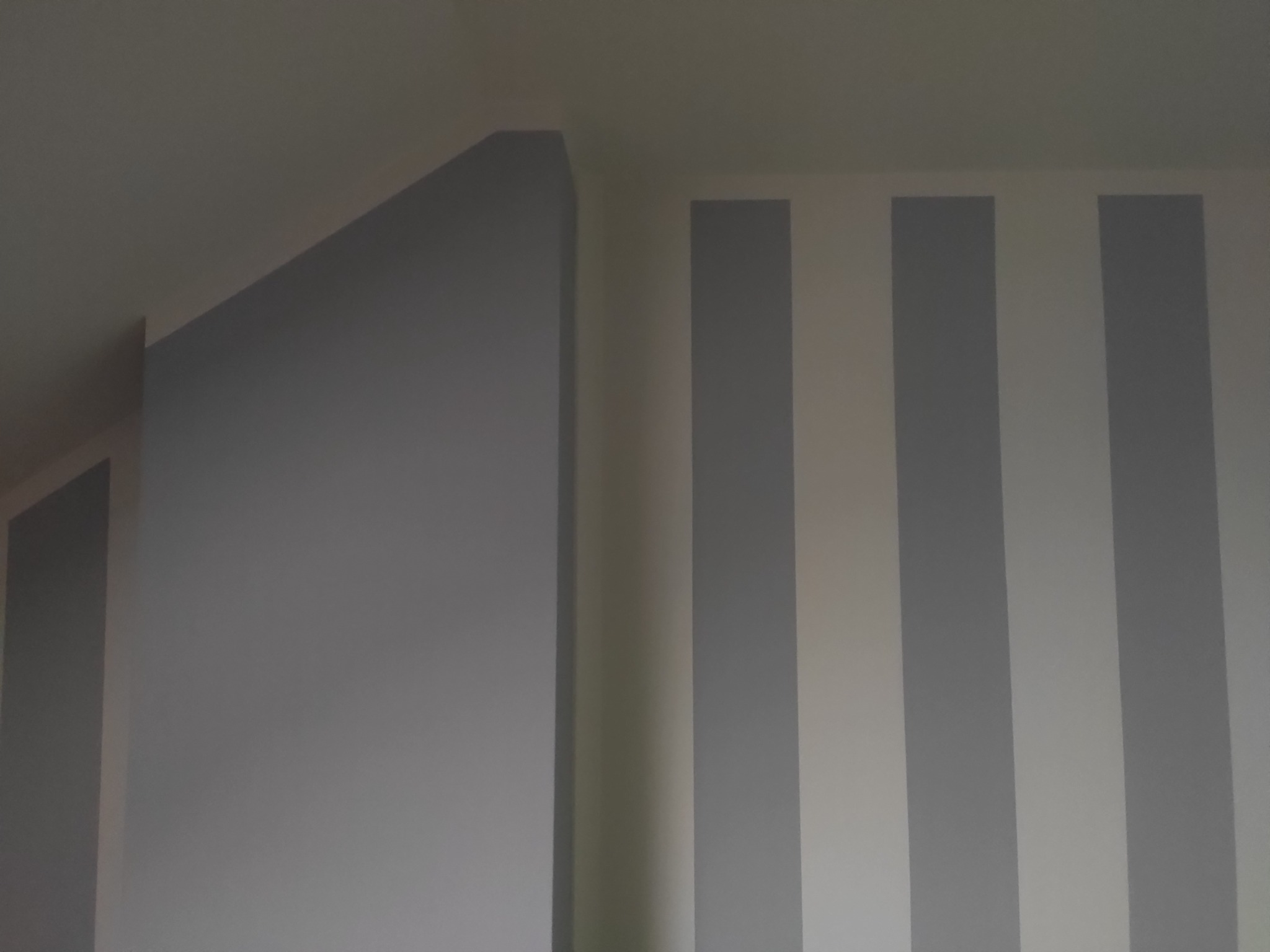 Pomieszczenie ze ścianami pomalowanymi w szaro-białe pionowe pasy oraz szarą ścianą pod skosem, widoczne połączenie ścian i sufitu.