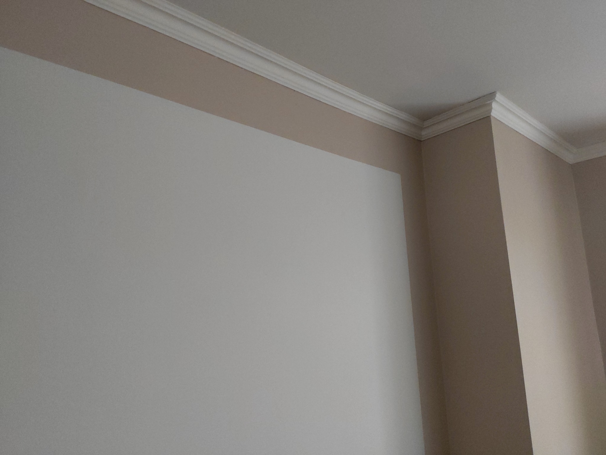 Pomieszczenie po remoncie: ściana w kolorze złamanej bieli z beżowym pasem pod sufitem i białym, ozdobnym gzymsem.