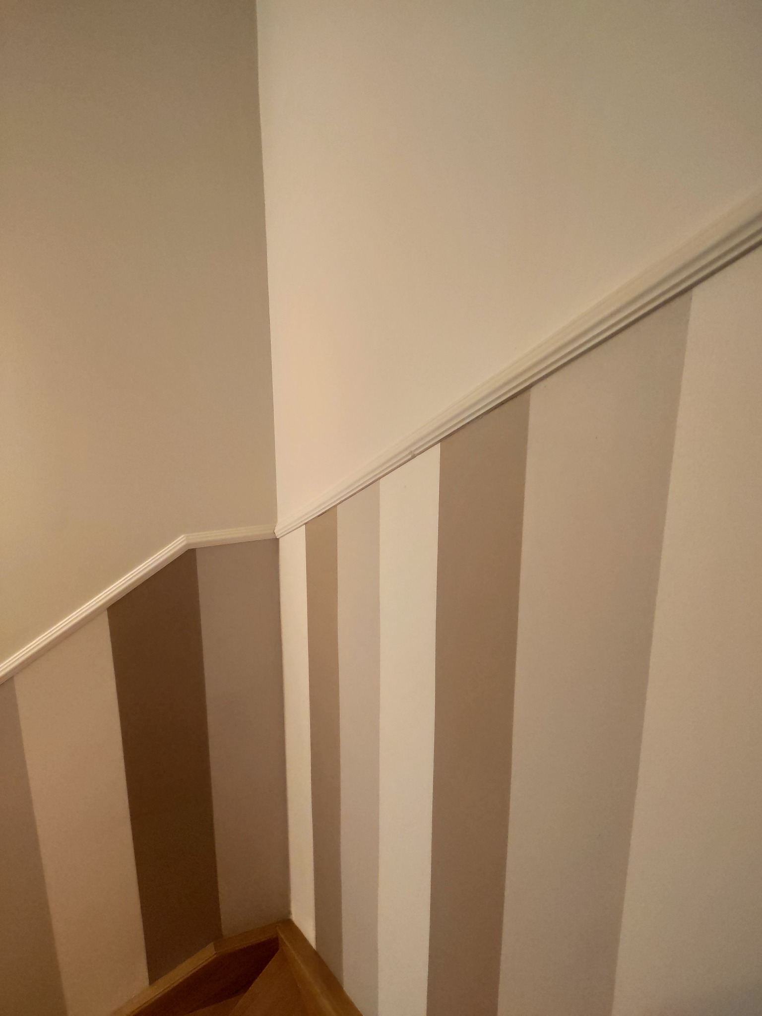 Ściana klatki schodowej z pionowymi pasami w odcieniach beżu i brązu, wykończona listwą przysufitową, widoczny fragment drewnianych schodów.