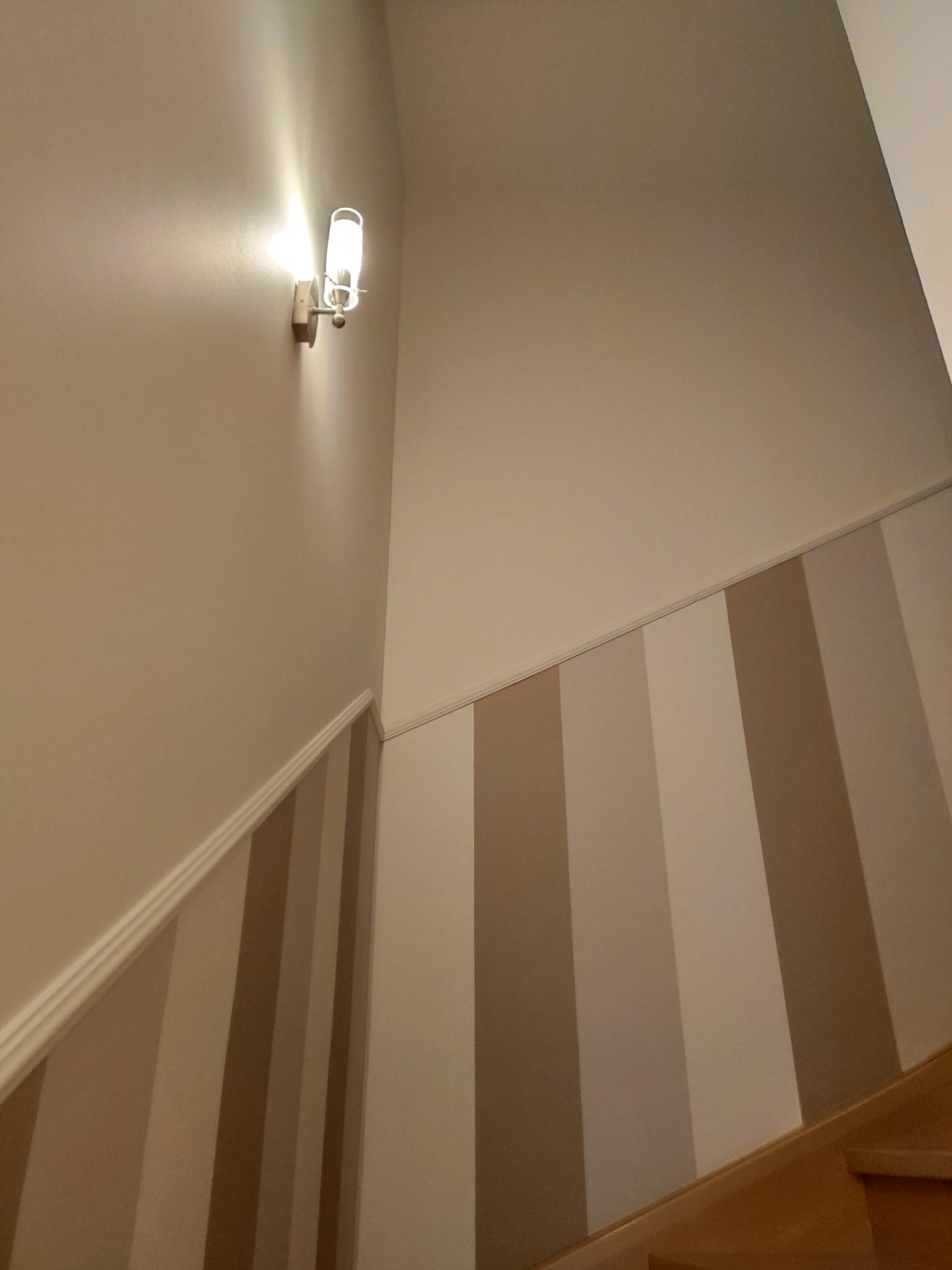 Klatka schodowa z paskami w odcieniach beżu i brązu, oświetlona kinkietem z żarówką w kształcie tuby.