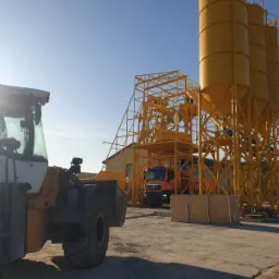 Widok na plac betoniarni z ładowarką kołową na pierwszym planie, pomarańczową ciężarówką MAN i żółtymi silosami na beton w tle, przy słonecznej pogodzie.
