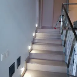 Jasne, drewniane schody z podświetleniem LED w ścianie, stalowa balustrada, widoczna drabina w tle.