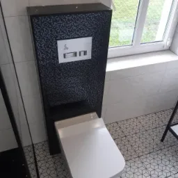 Podwieszana toaleta z czarną mozaiką na zabudowie stelaża, białe płytki ścienne i podłogowe z geometrycznym wzorem, widok z góry na fragment łazienki.