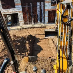 Widok z kabiny koparki na wykopany rów w ziemi, w tle budynek gospodarczy z drewnianymi bramami.