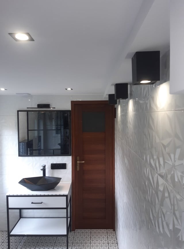 Nowoczesna łazienka z czarną, geometryczną umywalką nablatową, lustrem w czarnej ramie, drewnianymi drzwiami i trójwymiarowymi białymi płytkami na ścianie, oświetlona kwadratowymi lampami sufitowymi.