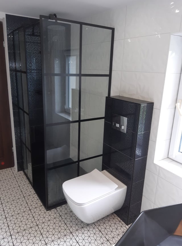 Nowoczesna łazienka z kabiną prysznicową typu walk-in z czarnymi profilami, podwieszaną miską WC z czarną obudową i geometryczną posadzką.