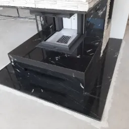 Częściowo obudowany kominek z czarnego marmuru, widoczne białe cegiełki na górze obudowy, surowy beton na podłodze.