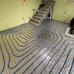 HYDRO POWER - Niebieskie rury ogrzewania podłogowego ułożone spiralnie na folii z czerwoną siatką, widoczne betonowe schody i surowa ściana z oznaczeniami w tle.