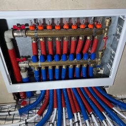 HYDRO POWER - Rozdzielacz ogrzewania podłogowego w metalowej szafce, z widocznymi czerwonymi i niebieskimi rurami doprowadzającymi, oraz rurami grzewczymi na folii izolacyjnej z rastrem.