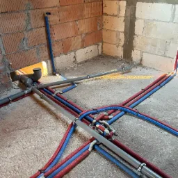 Surowy stan podłogi z rozłożonymi rurami instalacji wodnej i kanalizacyjnej w kolorach czerwonym, niebieskim i szarym, z widocznymi złączkami i obejmami, na tle surowych ścian z cegły i betonu...