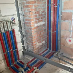 Instalacja wodna i kanalizacyjna w trakcie montażu: pionowe rury w kolorze czerwonym i niebieskim, mocowane do ściany z cegły i betonu komórkowego za pomocą metalowych uchwytów, oraz szare rury...