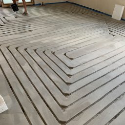 HYDRO POWER - Przygotowanie podłoża pod ogrzewanie podłogowe: wylewka z wyfrezowanymi kanałami na rury, widoczna osoba z wiadrem i fragment instalacji.