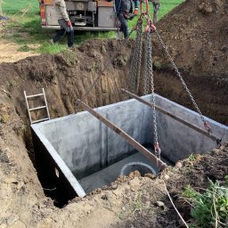 HYDRO POWER - Montaż betonowego zbiornika podziemnego z użyciem dźwigu na zewnątrz, widoczni dwaj pracownicy w odzieży roboczej, drabina oparta o wykop