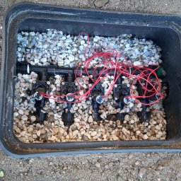 HYDRO POWER - Zawory nawadniające ukryte w plastikowej skrzynce wypełnionej ozdobnymi kamieniami, z widocznymi czerwonymi przewodami sterującymi.