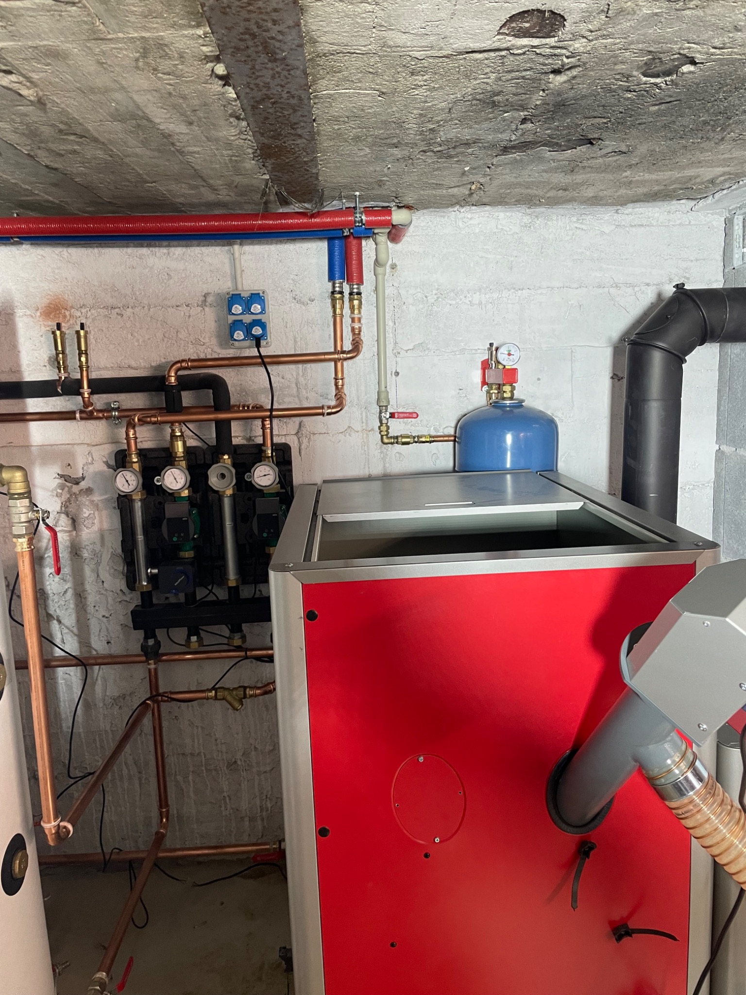 Nowoczesny piec CO z czerwonym panelem, podłączony do miedzianej instalacji grzewczej z manometrami, zamontowany w pomieszczeniu z surowym betonowym stropem.
