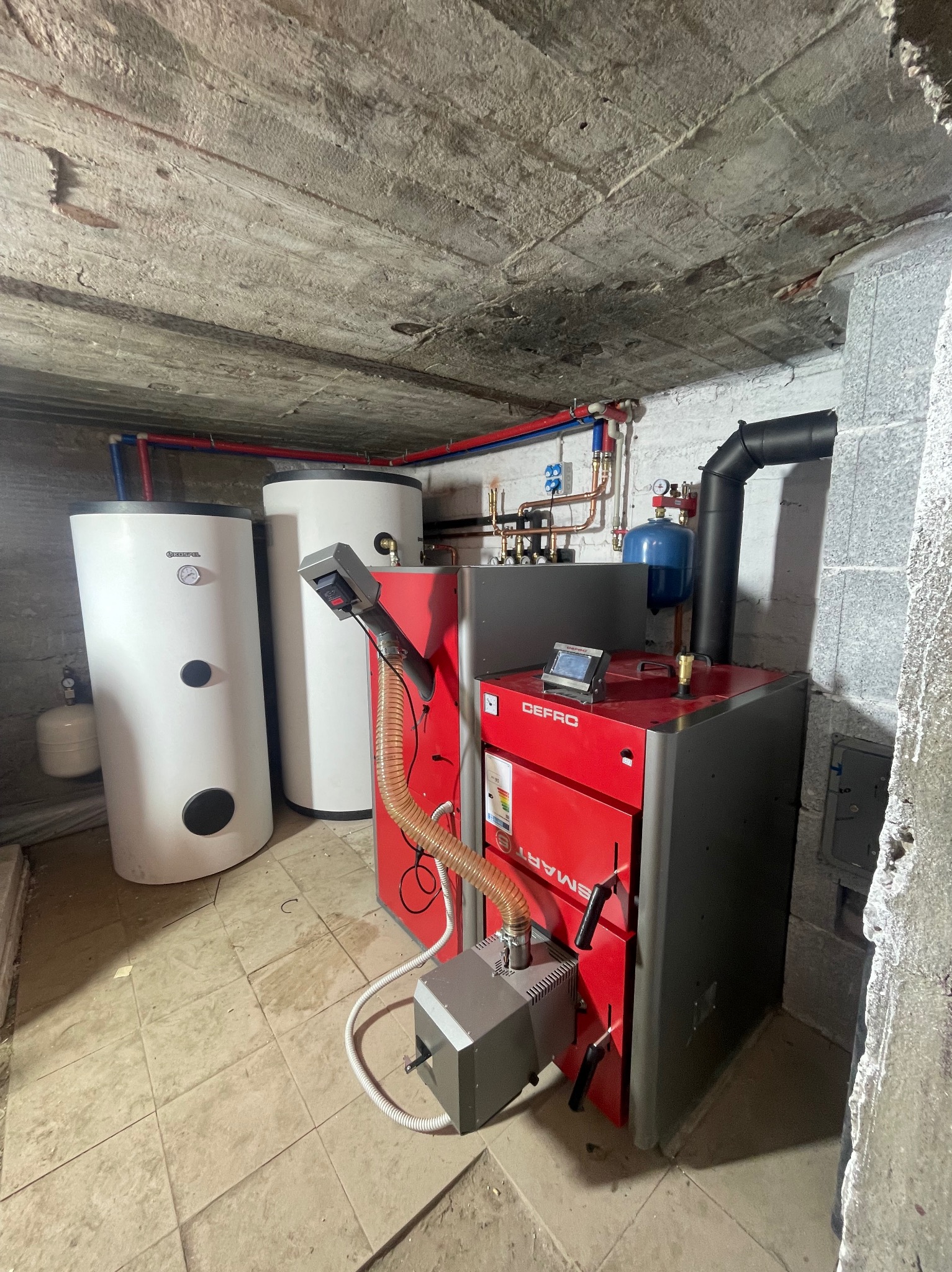 Czerwono-szary piec CO marki Defro Smart z podajnikiem, dwa białe zasobniki ciepłej wody użytkowej Kospel i instalacja grzewcza w pomieszczeniu z surowym betonowym sufitem.