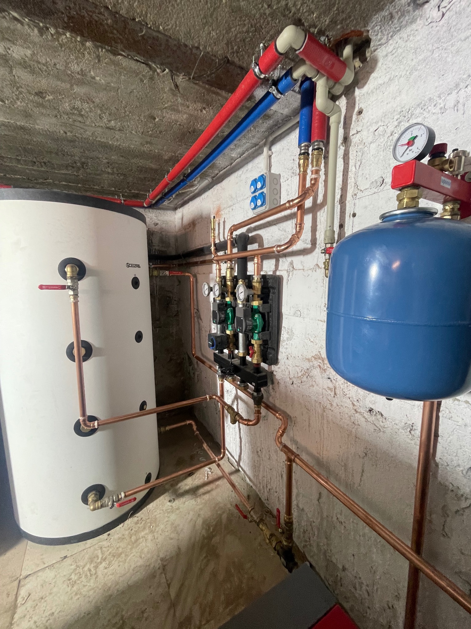 Nowoczesny system instalacji grzewczej z białym zasobnikiem ciepłej wody, niebieskim naczyniem wzbiorczym i miedzianymi rurami rozprowadzonymi na tle surowej, pomalowanej ściany piwnicy.
