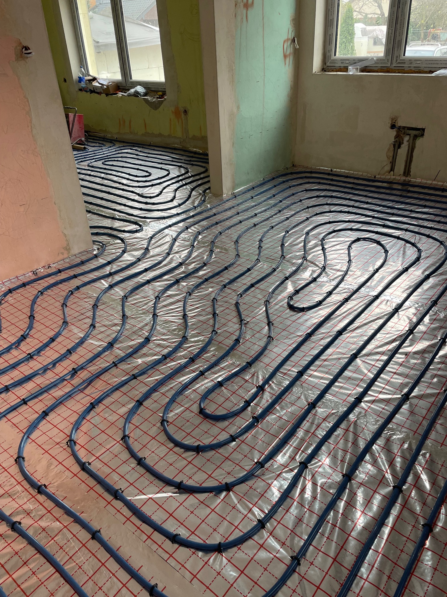 Niebieskie rury ogrzewania podłogowego ułożone na srebrnej folii z czerwoną siatką w surowym wnętrzu z oknami i widocznymi śladami po pracach budowlanych.