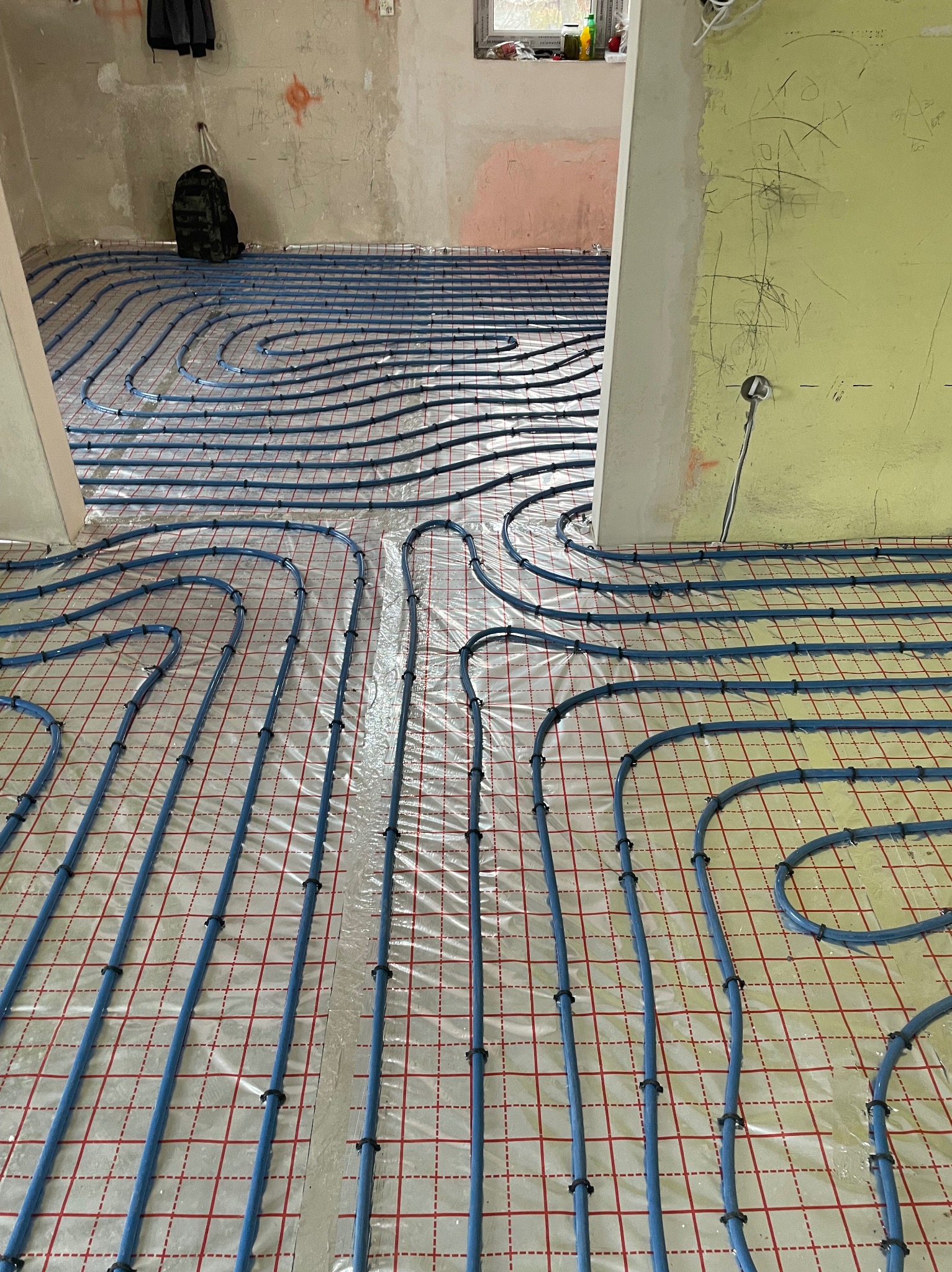 Niebieskie rury systemu ogrzewania podłogowego ułożone na folii z czerwoną siatką, widoczne surowe ściany w trakcie remontu, plecak w tle.