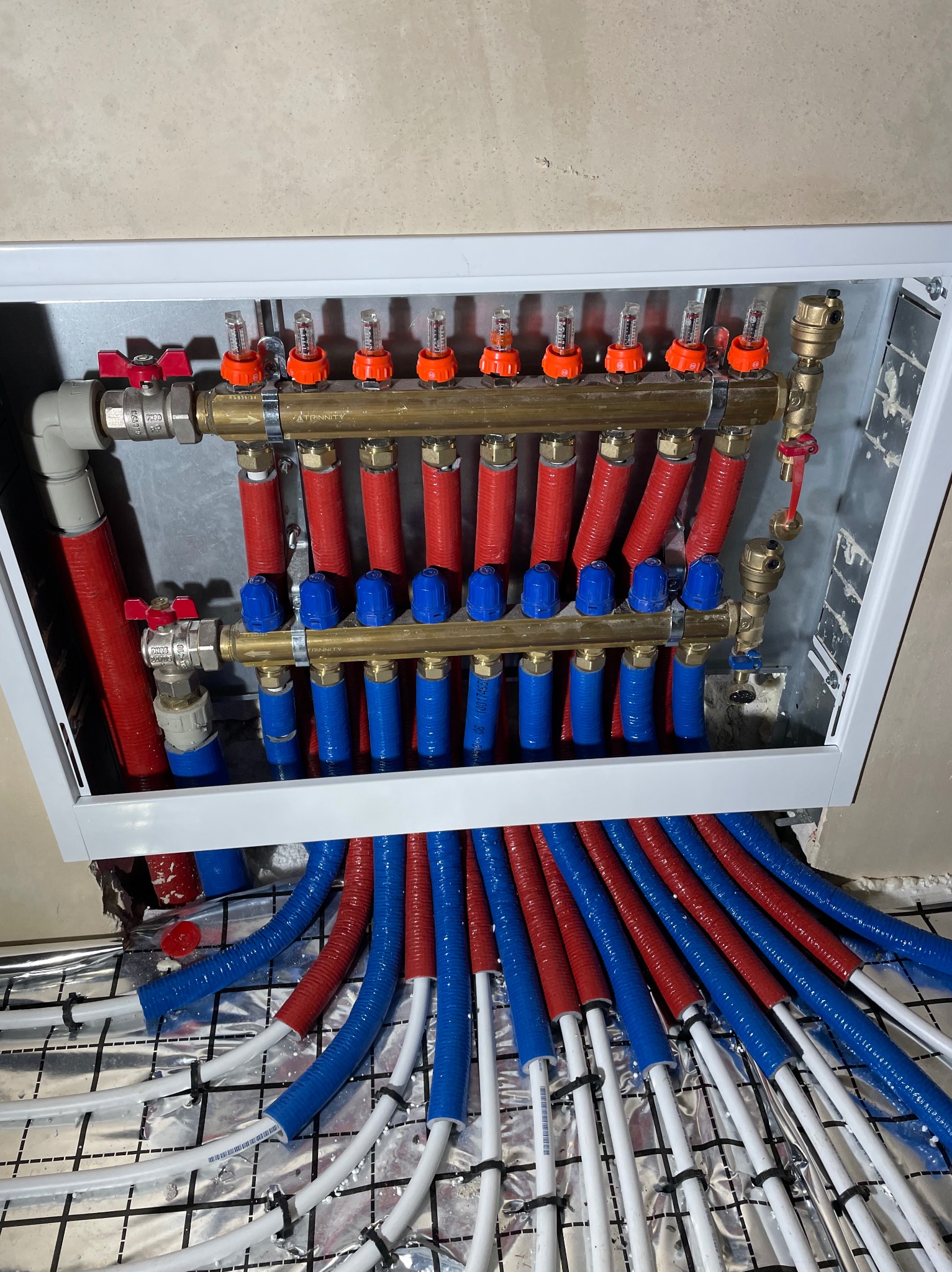 Rozdzielacz ogrzewania podłogowego w metalowej szafce, z widocznymi czerwonymi i niebieskimi rurami doprowadzającymi, oraz rurami grzewczymi na folii izolacyjnej z rastrem.