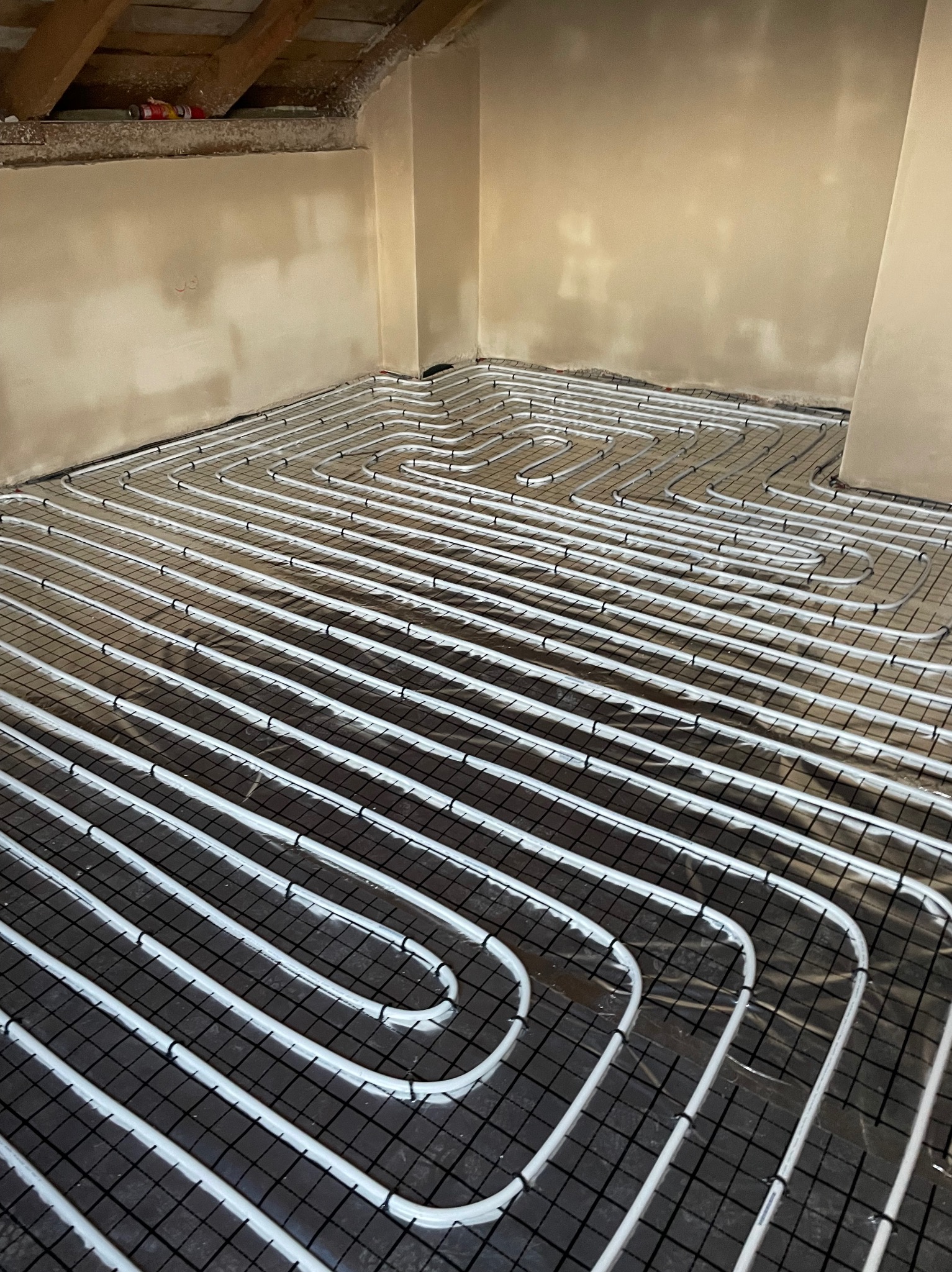 Białe rury grzewcze ułożone w pętle na czarnej siatce podłogowej w surowym wnętrzu z otynkowanymi ścianami, przygotowane do wylania posadzki.
