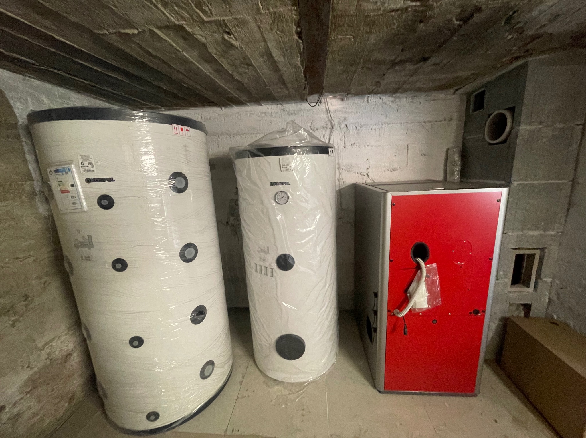 Nowe, owinięte folią bąbelkową zbiorniki ciepłej wody użytkowej marki Kospel oraz piec CO z czerwonym panelem frontowym, ustawione w piwnicy o surowych ścianach z drewnianym belkowaniem stropu.