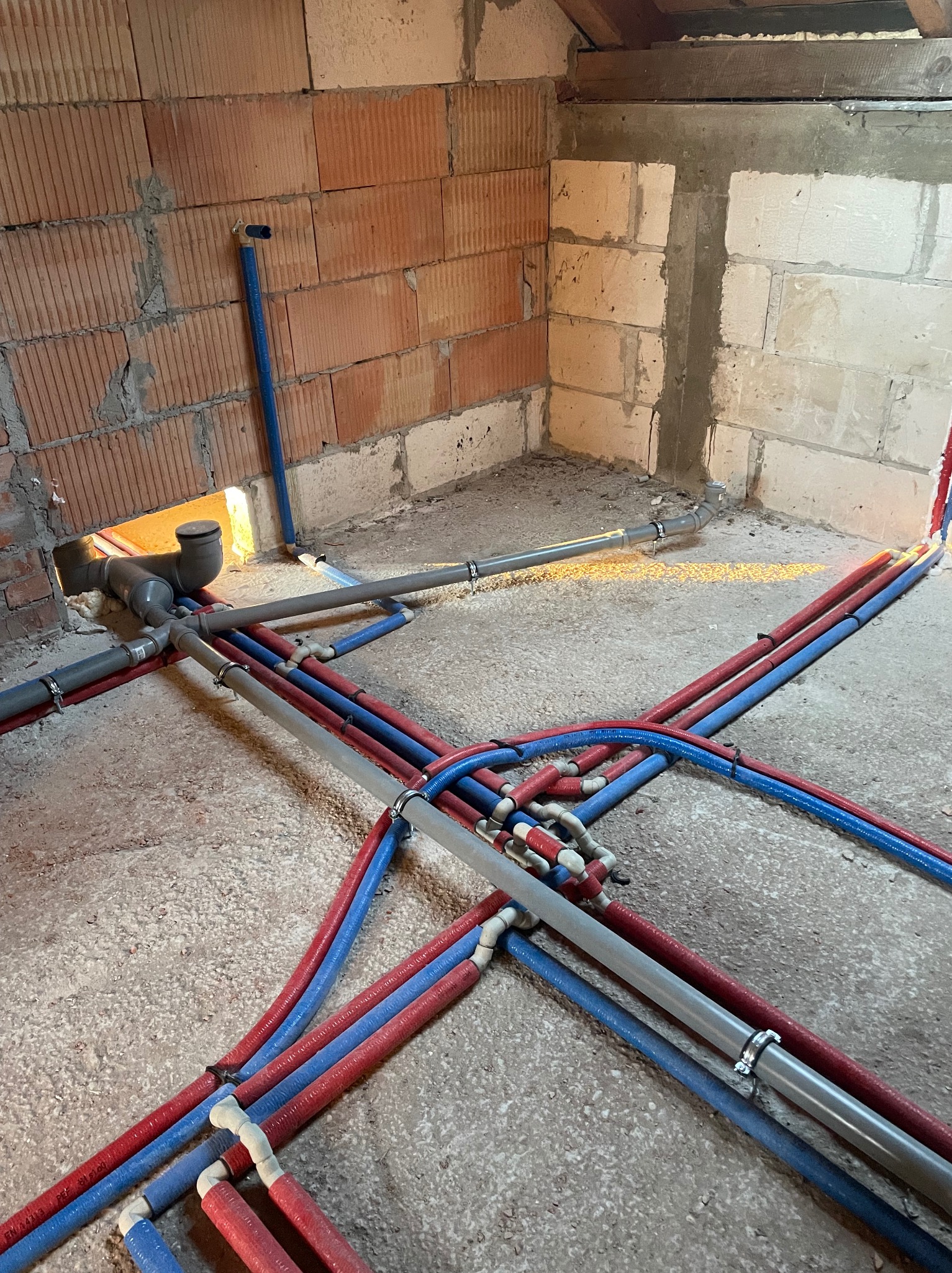 Surowy stan podłogi z rozłożonymi rurami instalacji wodnej i kanalizacyjnej w kolorach czerwonym, niebieskim i szarym, z widocznymi złączkami i obejmami, na tle surowych ścian z cegły i betonu...