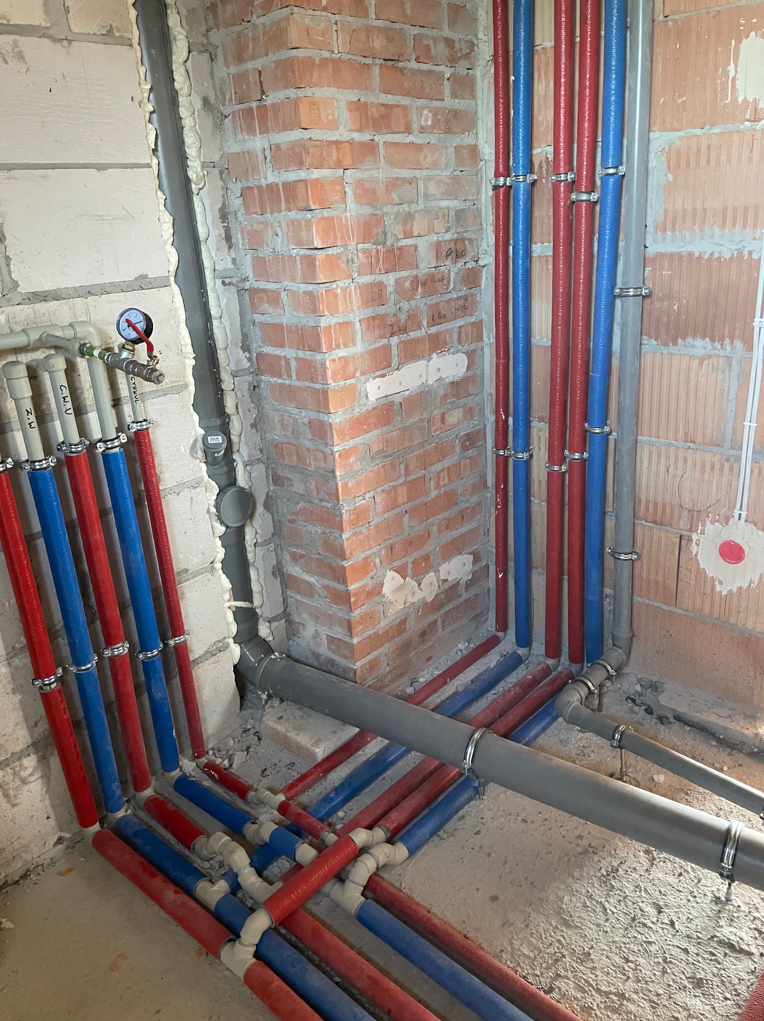 Instalacja wodna i kanalizacyjna w trakcie montażu: pionowe rury w kolorze czerwonym i niebieskim, mocowane do ściany z cegły i betonu komórkowego za pomocą metalowych uchwytów, oraz szare rury...