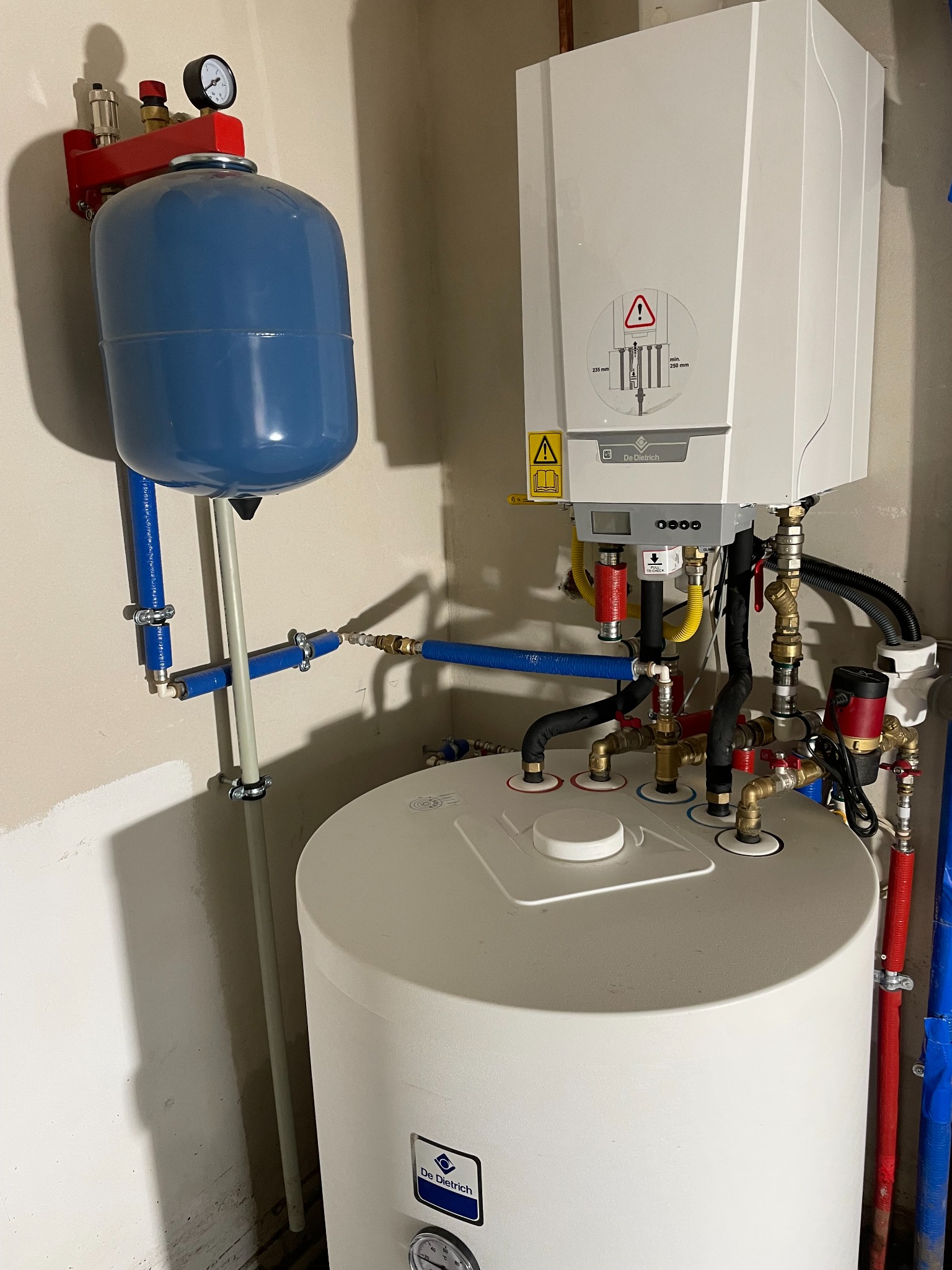 Nowoczesny, biały piec gazowy De Dietrich z podłączonymi rurami (czerwone, niebieskie, czarne) i zbiornikiem wyrównawczym. Manometr na zbiorniku. Ściana w tle.