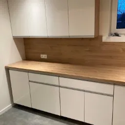 Nowoczesna zabudowa kuchenna z białymi frontami i drewnianym blatem, widoczna część ściany z gniazdkami elektrycznymi i fragment okna.