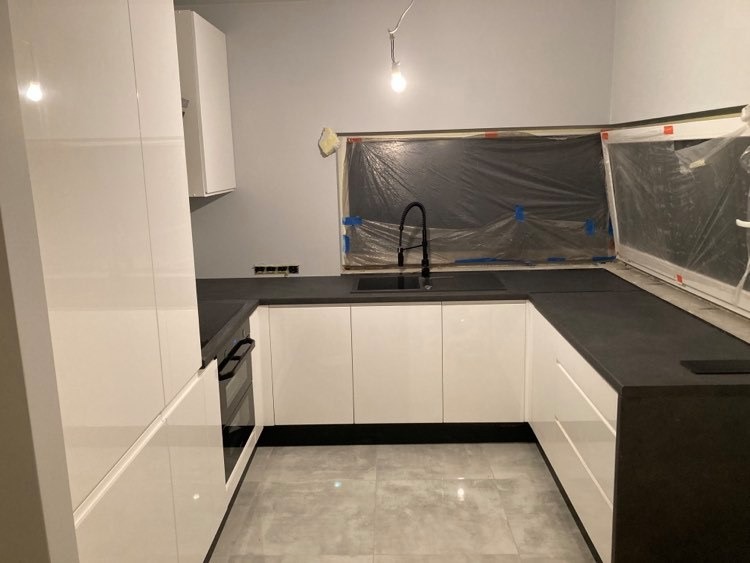 Nowoczesna kuchnia w trakcie montażu: białe, lakierowane szafki z czarnym blatem, okno zabezpieczone folią malarską, widoczne gniazdka elektryczne.