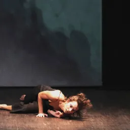 Kobieta w czarnym stroju leży na drewnianej scenie, wykonując ekspresyjny ruch taneczny na tle ciemnego, abstrakcyjnego tła.
