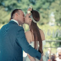 Pocałunek pary młodej w plenerze, panna młoda z kwiatowym wiankiem i odkrytymi ramionami, pan młody w niebieskim garniturze, rozmyte tło z zielenią.