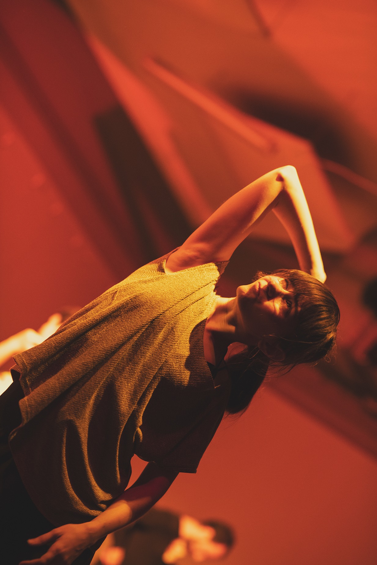 Kobieta w koszulce z krótkim rękawem, z podniesioną ręką, w ciepłym, pomarańczowym oświetleniu scenicznym, na tle geometrycznych świateł.