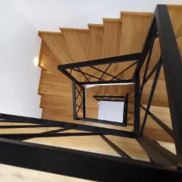 Spiralne schody drewniane z czarną metalową balustradą, widziane z góry, z minimalistycznym białym tłem.