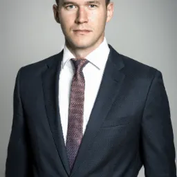 Portret młodego mężczyzny w ciemnym garniturze, białej koszuli i wzorzystym krawacie, na jednolitym jasnoszarym tle.