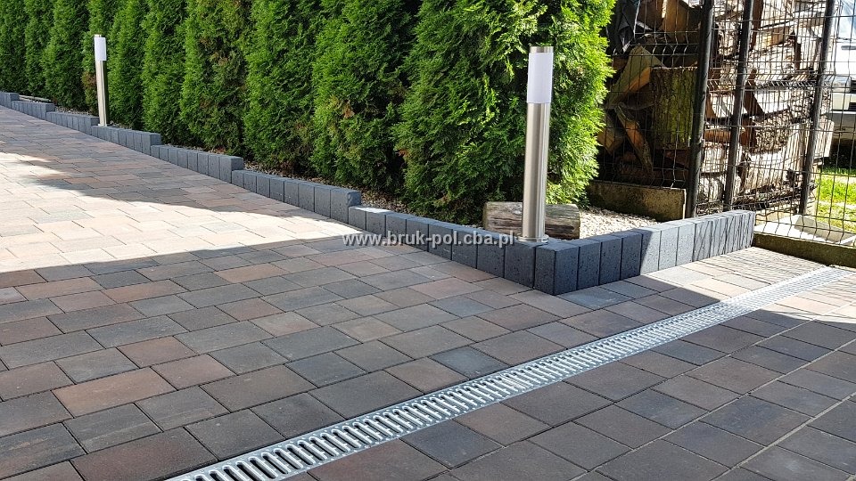 Ułożona kostka brukowa w Tucholi z odpływem liniowym, obrzeżami i lampami ogrodowymi, w tle zielone tuje i drewno składowane w metalowej siatce.