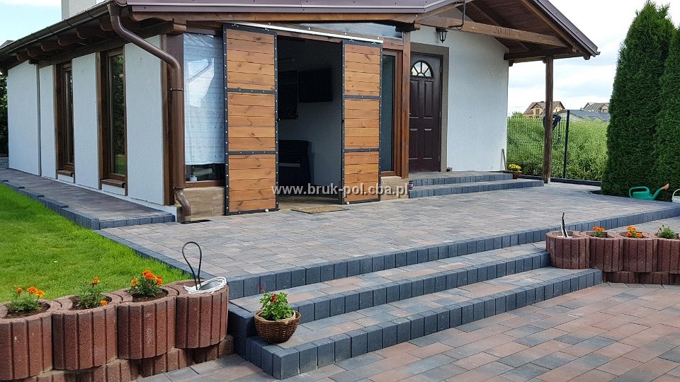 Nowoczesny taras z kostki brukowej w odcieniach szarości i czerwieni, z niskimi schodami i dekoracyjnymi donicami z kwiatami, na tle białego domu z drewnianymi elementami i przesuwnymi drzwiami.