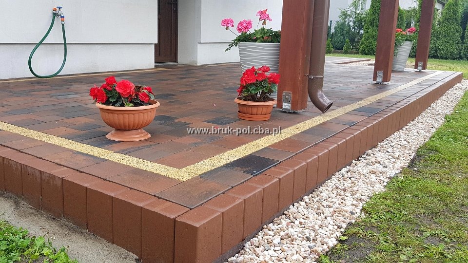 Wykończony taras z wielobarwnej kostki, obrzeża z brązowych bloczków, oddzielone od trawnika białym żwirem, donice z czerwonymi kwiatami, widoczny kran z wężem na ścianie budynku.