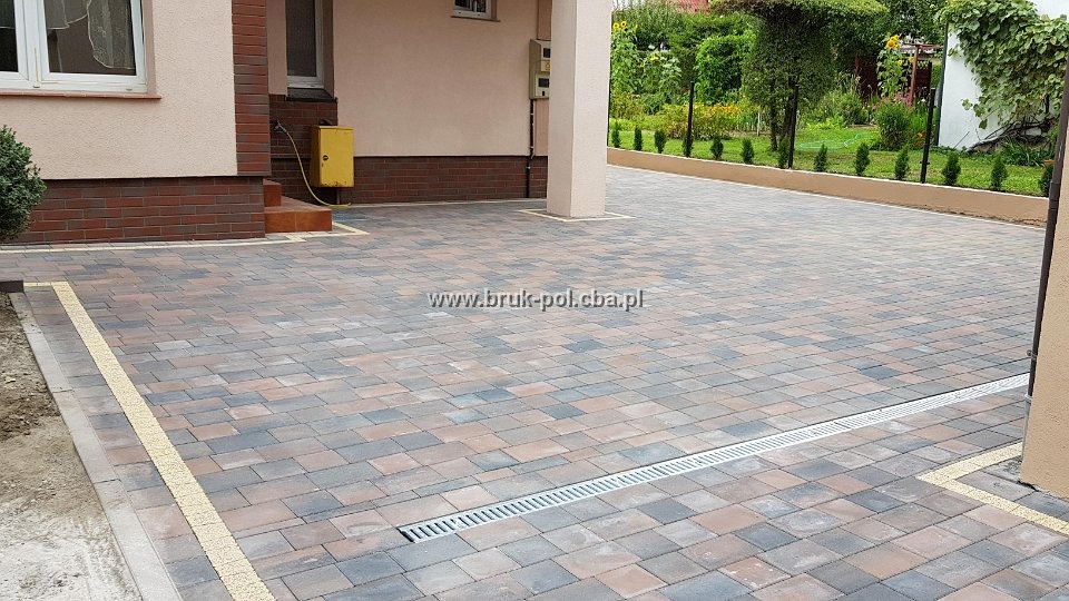 Ułożona kostka brukowa w odcieniach brązu i szarości, z widocznym odpływem liniowym i żółtą linią oddzielającą od trawnika, na tle domu z kolumną.