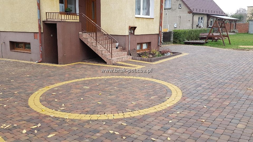 Ułożona kostka brukowa przed domem z żółtym okręgiem i obrzeżami, widoczne schody wejściowe i fragment ogrodu.