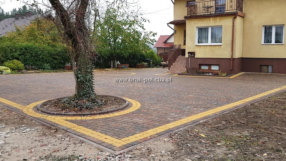 Rozległy podjazd z kostki brukowej w odcieniach brązu, obramowany żółtą kostką, okalający drzewo z bluszczem, na tle dwukondygnacyjnego domu z balkonem.