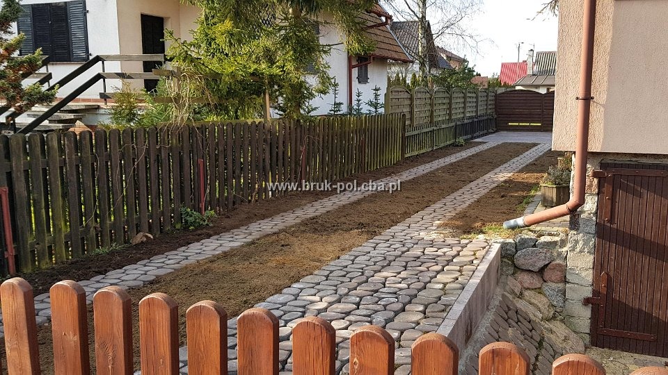 Ułożona z okrągłych kamieni ścieżka prowadząca do posesji, widok zza drewnianego płotku, świeżo obsypana ziemią po bokach.