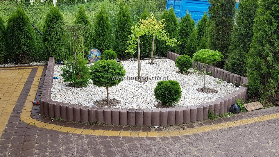 Obrzeże rabaty z brązowych betonowych palisad, wypełnione białym żwirem i ozdobnymi drzewkami, otoczone tujami i kostką brukową w odcieniach brązu i żółci.