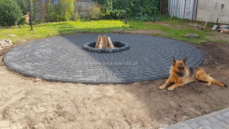 Okrągły plac z szarej kostki brukowej z paleniskiem pośrodku, na pierwszym planie leży owczarek niemiecki, w tle trawnik, krzewy i ogrodzenie.