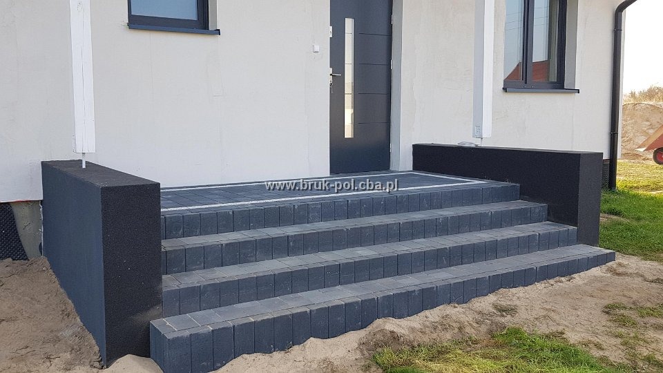Nowoczesne schody wejściowe z ciemnoszarej kostki brukowej, wykończone czarnymi blokami po bokach, prowadzące do szarych drzwi wejściowych domu z białymi ścianami.