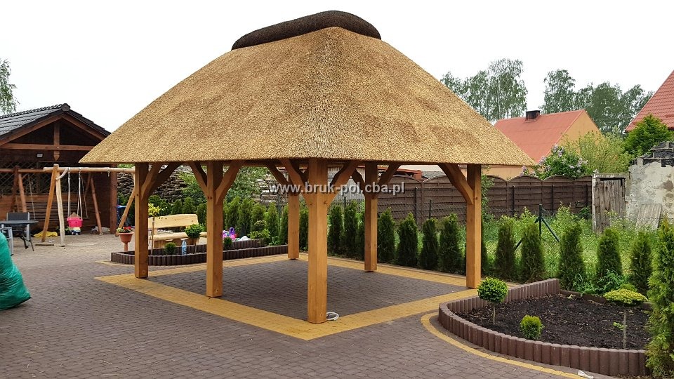Zadaszona altana z drewnianą konstrukcją i strzechą, otoczona brązowo-żółtą kostką brukową, z widocznymi elementami ogrodu i roślinnością.