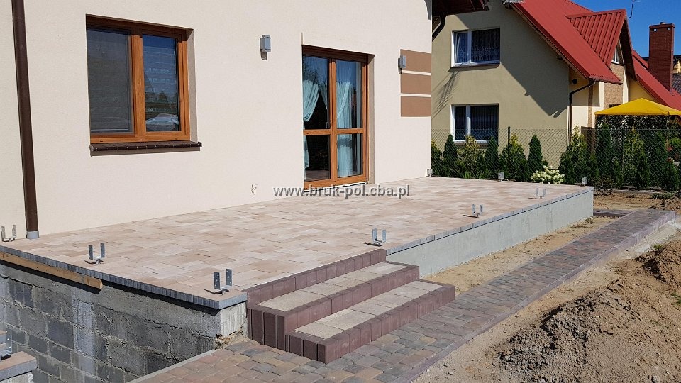 Wykończony taras z kostki brukowej w odcieniach beżu i brązu, z trzema schodami prowadzącymi do drzwi tarasowych, widoczne metalowe wsporniki i fragment ścieżki z kostki.